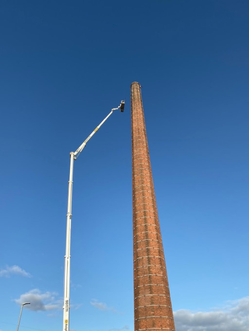 Looking up at tall brick chimney from the ground as a white cherrypicker expands to scale it against a blue sky.