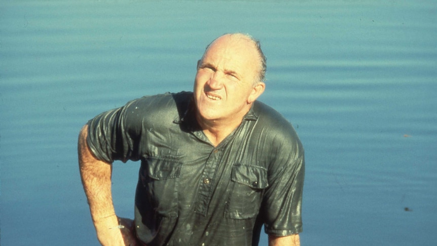 A man in a safari shirt is standing in still water and completely drenched.