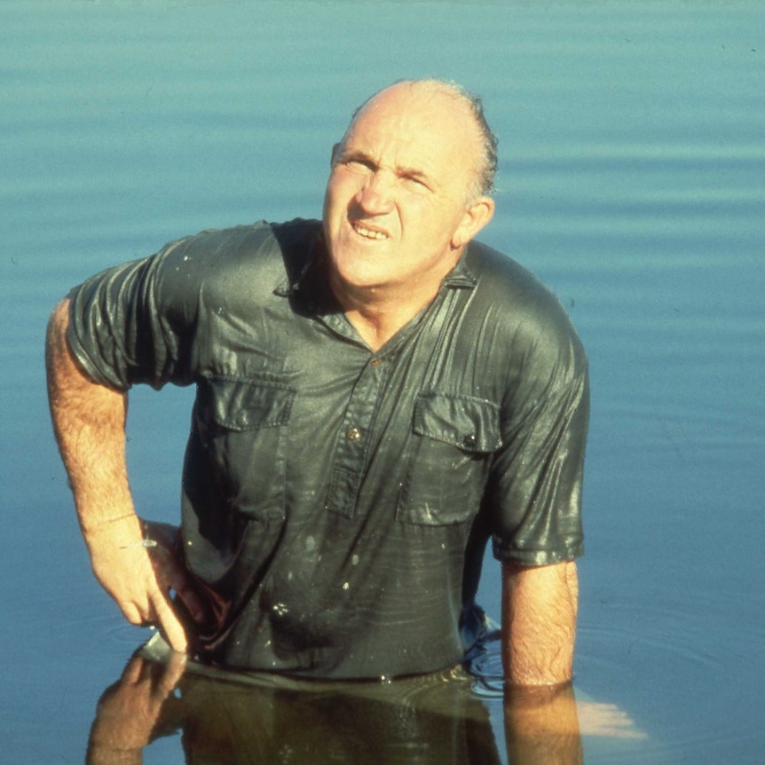 A man in a safari shirt is standing in still water and completely drenched.