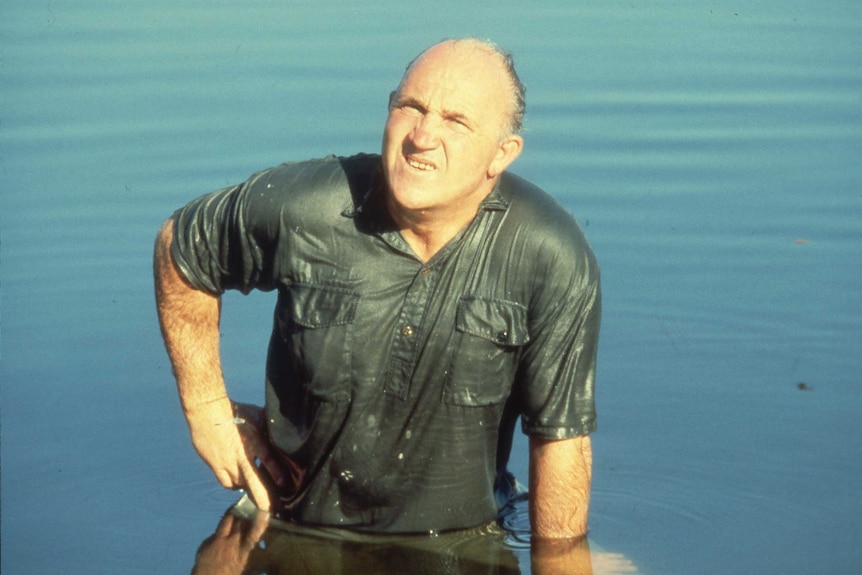 A wet man stands in a body of water, hand on one hip.