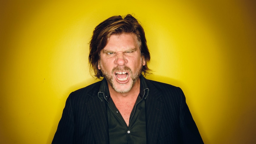 Tex Perkins stands against a yellow backdrop