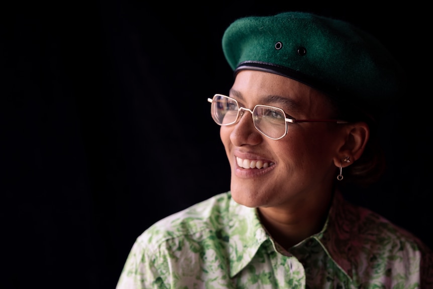 A woman wears a deep green beret and a green printed shirt. She is smiling slightly and looking away from the camera.