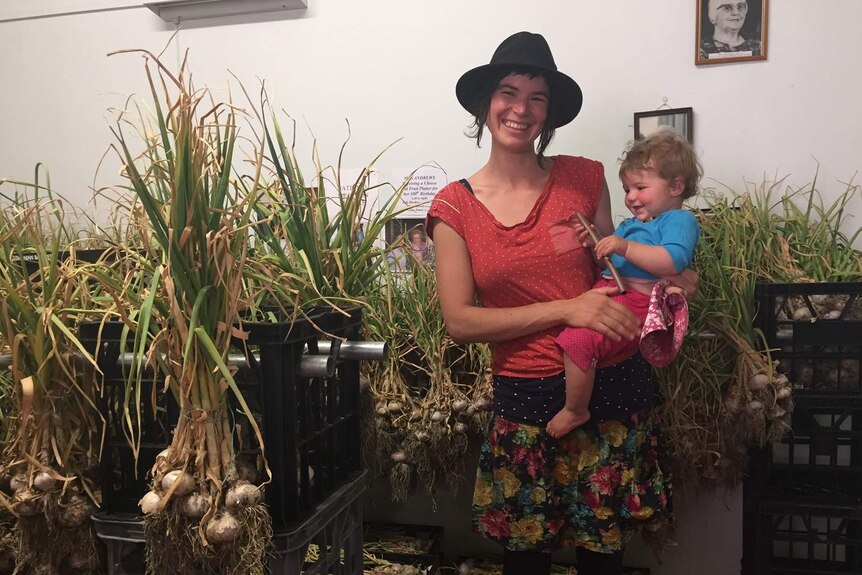 A young woman, with a baby on her hip, stands beside harvested garlic.