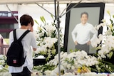 A young man in white T-shirt prays next to white flowers and memorial photo of Shinzo Abe