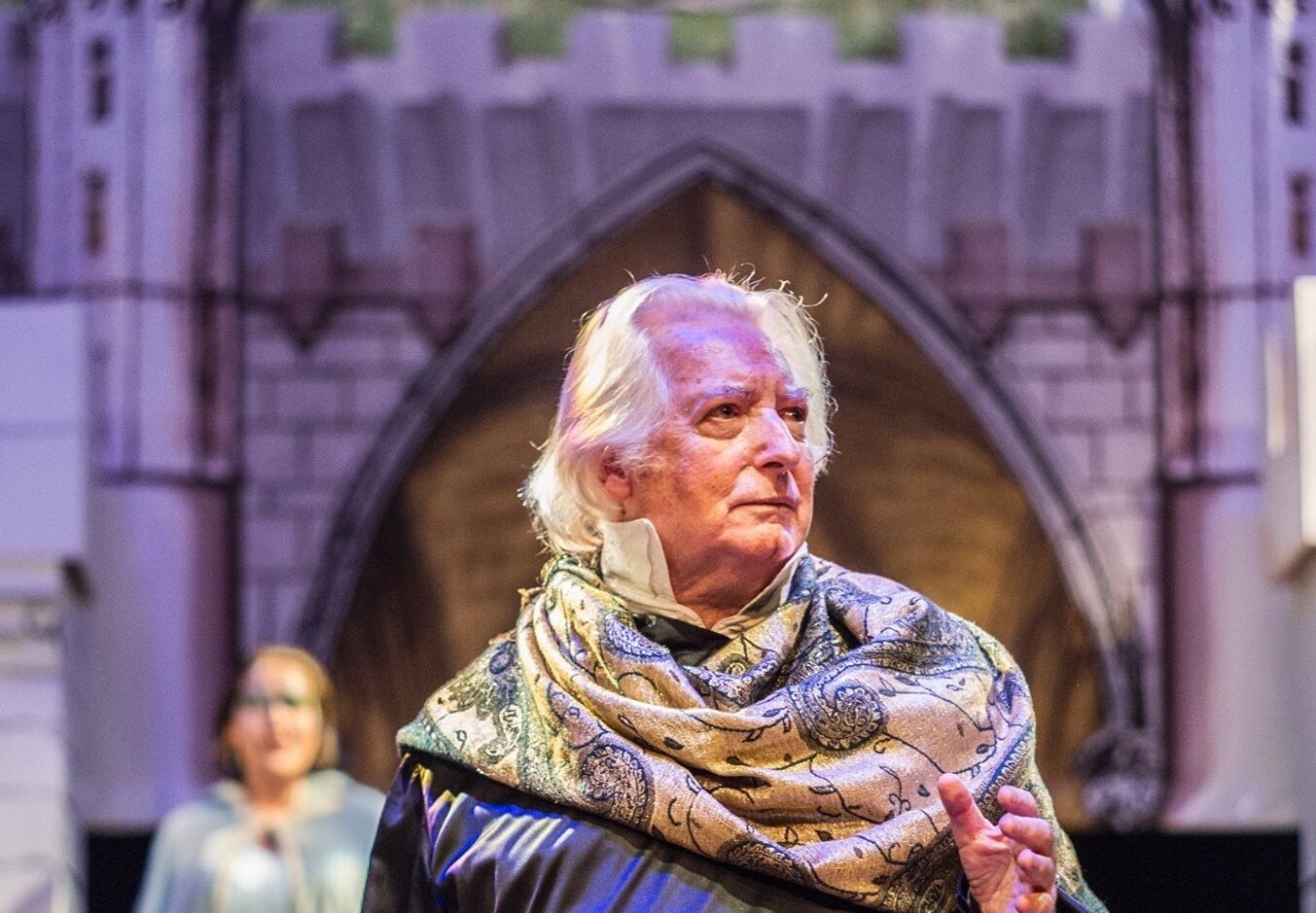 A white haired man stands on a stage with a scarf draped around his shoulders