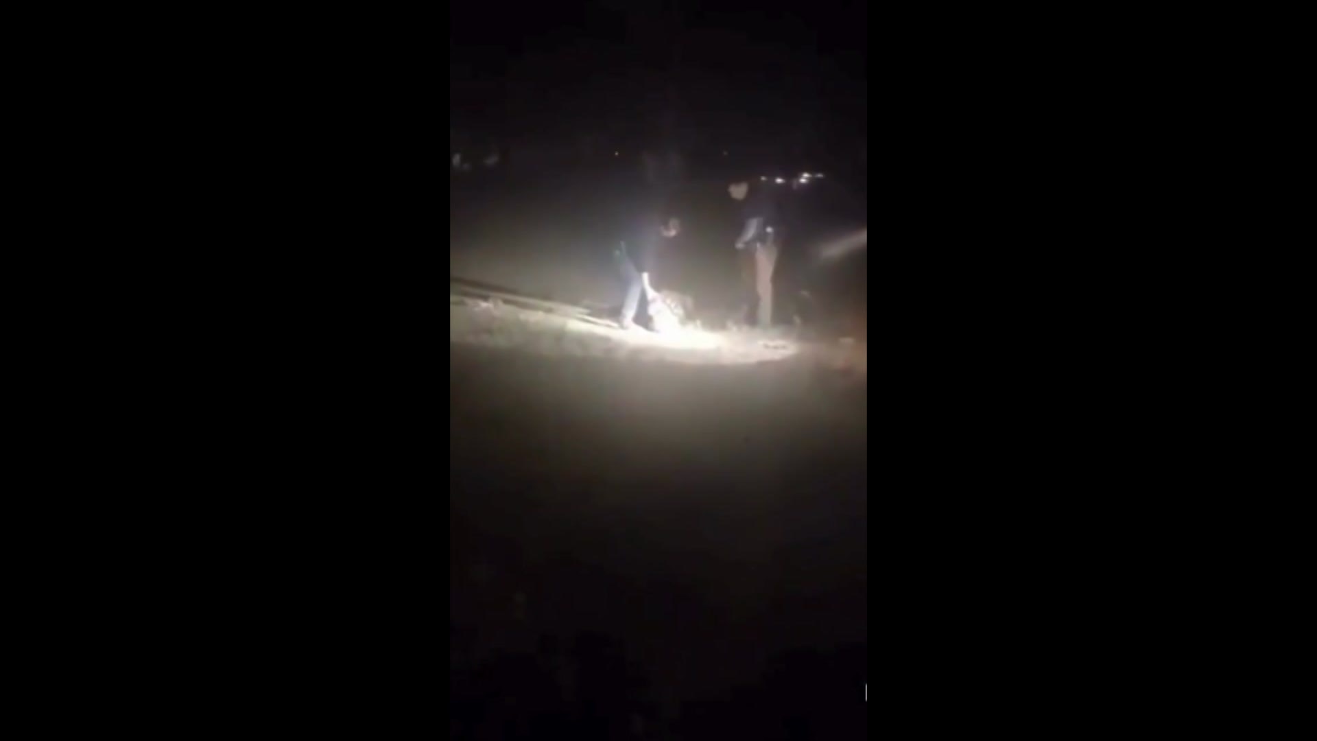 A dark grainy image of two officers leaning down to hold someone laying on the ground, with a white spotlight on them.