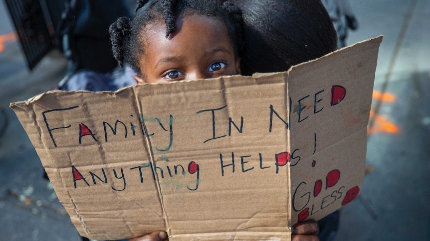 A toddler holds a sign over her face that reads "family in need"