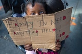 A toddler holds a sign over her face that reads "family in need"