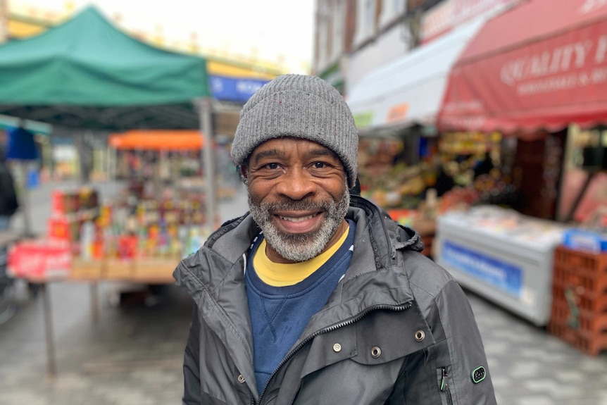A man wearing a jacket and a beanie smiles, he is at a market.