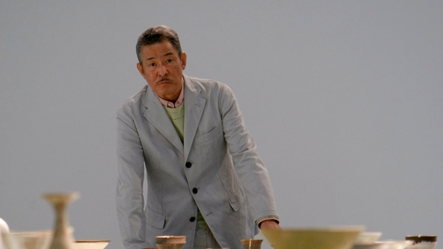 A Japanese man in a light suit stands in front of a table topped with pottery