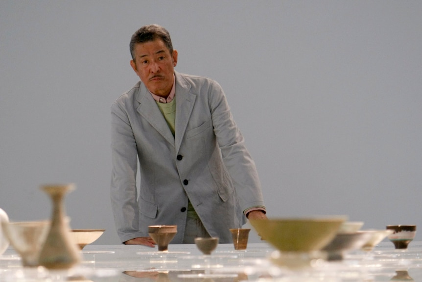 A Japanese man in a light suit stands in front of a table topped with pottery