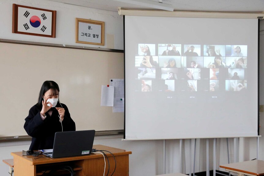 A teacher wearing a mask gives the OK symbol to a laptop in an empty classroom. A video conference screen shows students' faces
