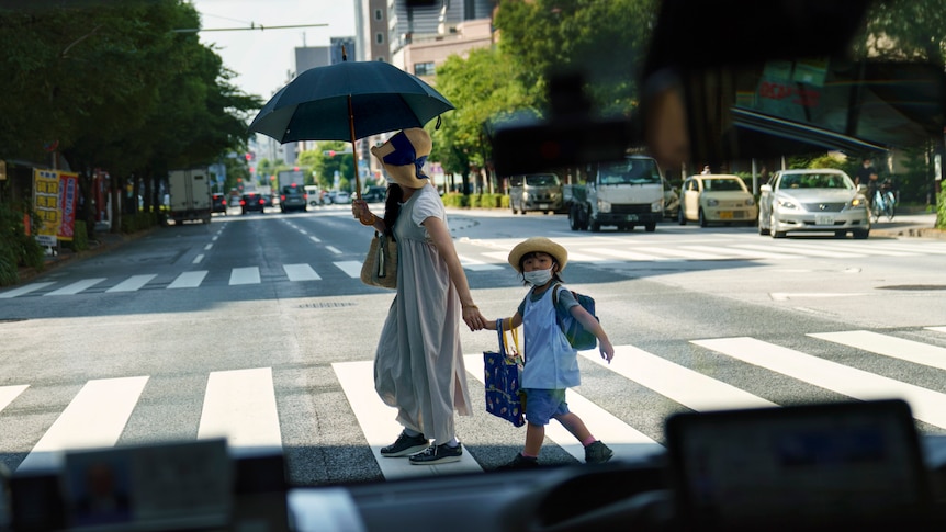 mother holds hand of child as they cross the road holding an umbrella. 