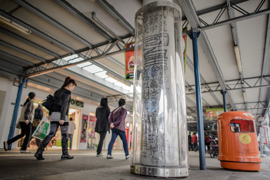 A pillar painted with black calligraphy shielded beneath a surrounding of clear plastic.
