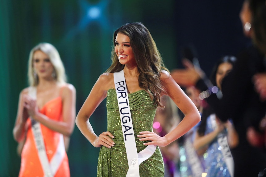 A woman in sparkly green dress, with a white sash saying PORTUGAL, stands smiling with hands on hips, on stage
