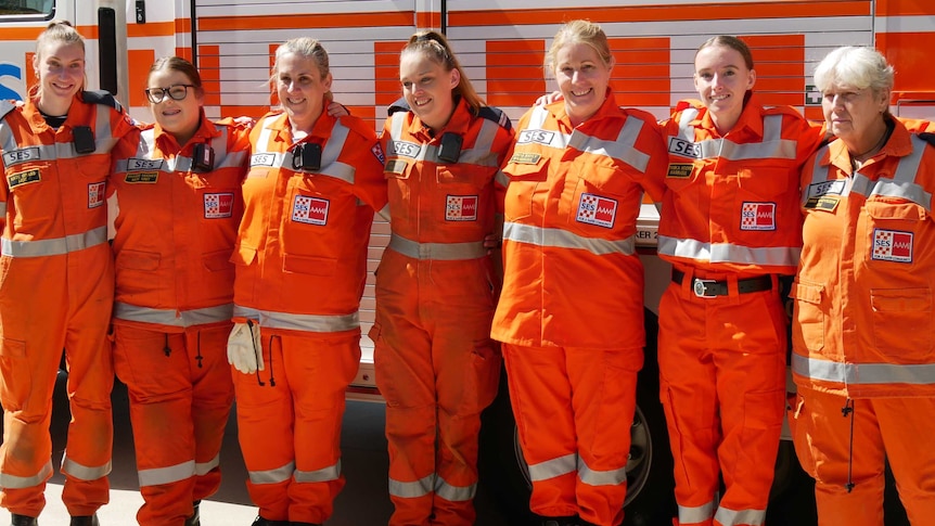 Seven smiling women in SES orange jumpsuits stand arm in arm in front of an Emergency Service truck.