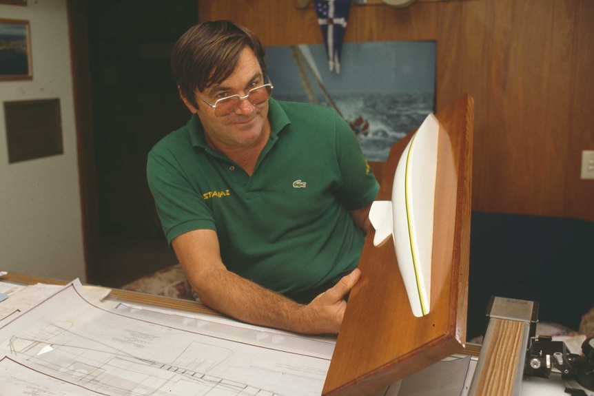 A smiling, dark-haired man in glasses displays a small replica of a yacht.
