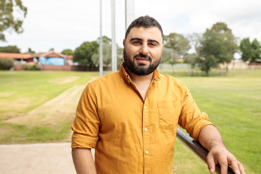 Young man with dark beard and moustache, wearing turmeric-coloured button down shirt, standing, with sports oval in background.