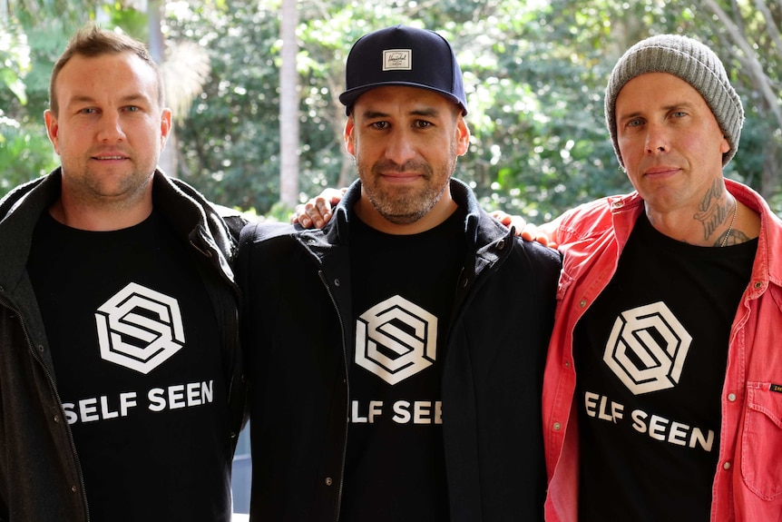 Three men standing with their arms around each other wearing a black T for their group Self Seen.