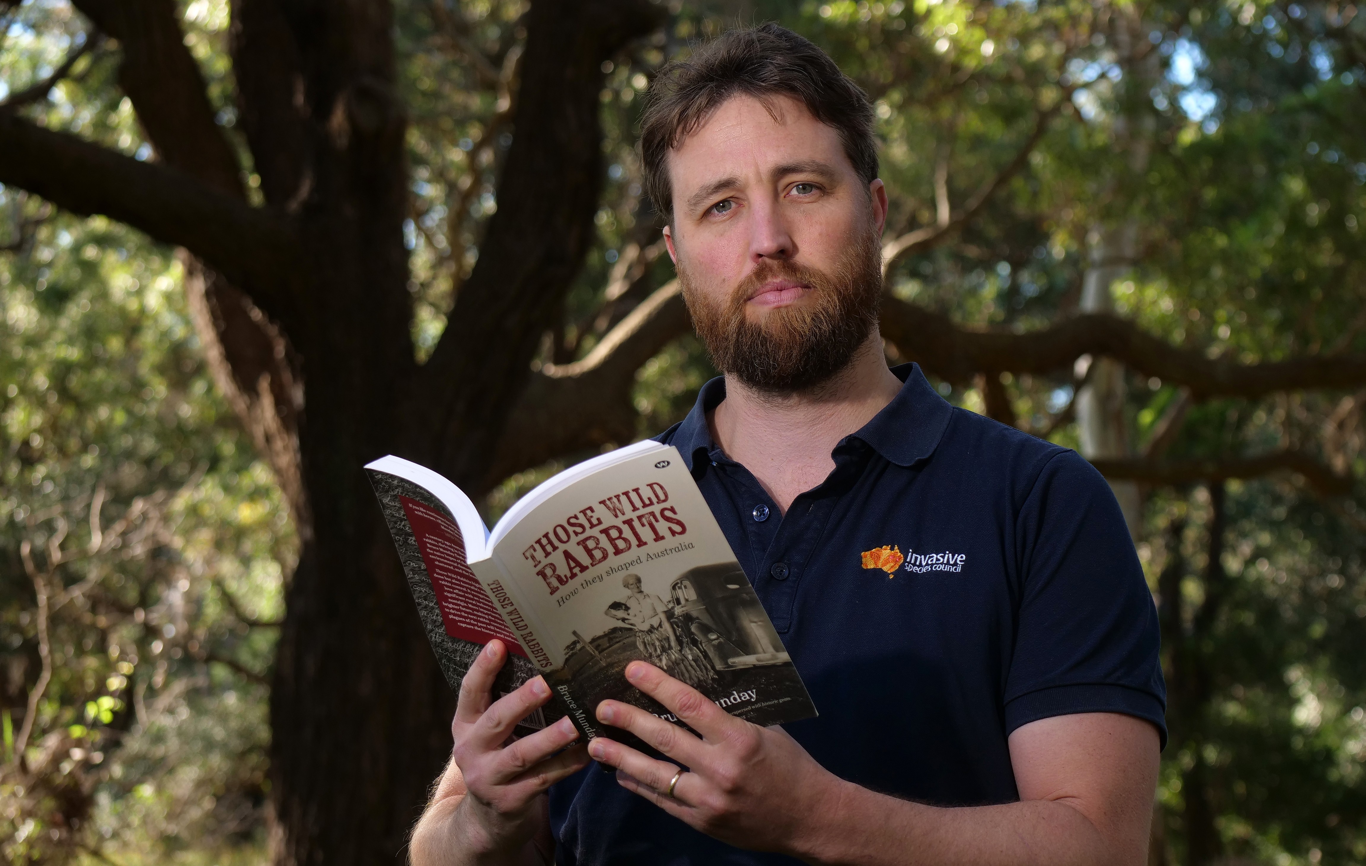 A fair-skinned, brown-haired bearded man, Jack, stands under a tree holding a book that says 'Those Wild Rabbits'.