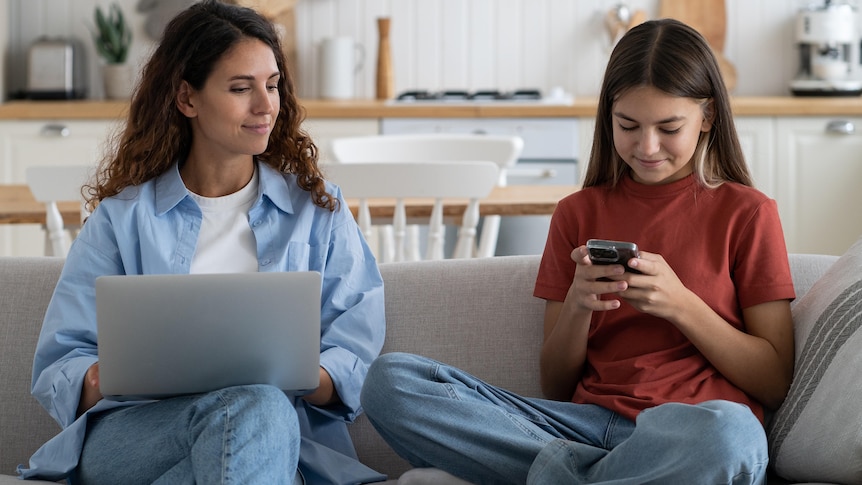 A curious mother sits beside her daughter on a couch and spies on her phone use.