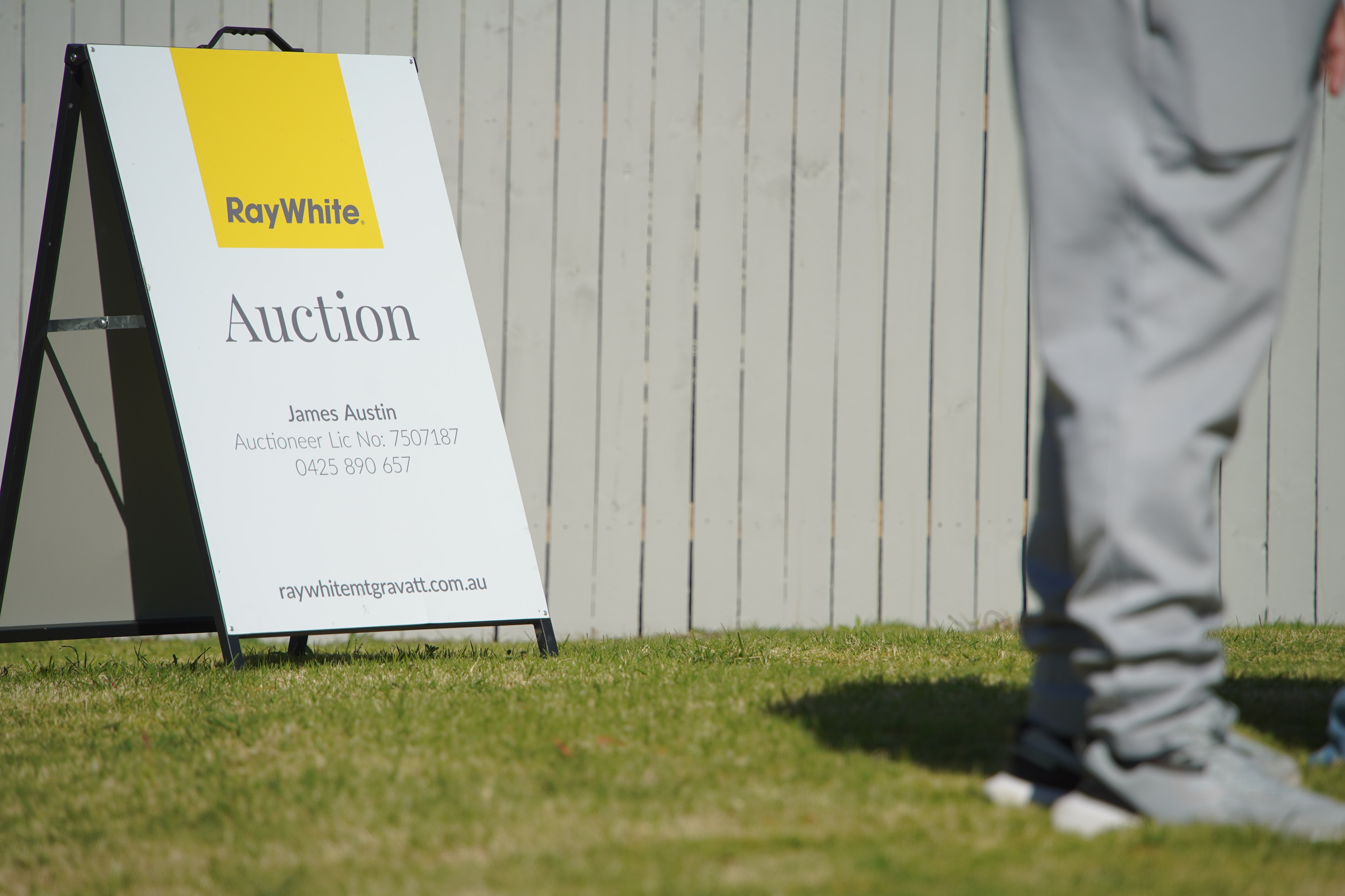 An A-frame sign which reads 'Auction' sitting on a patch of grass in front of a wooden picket fence.