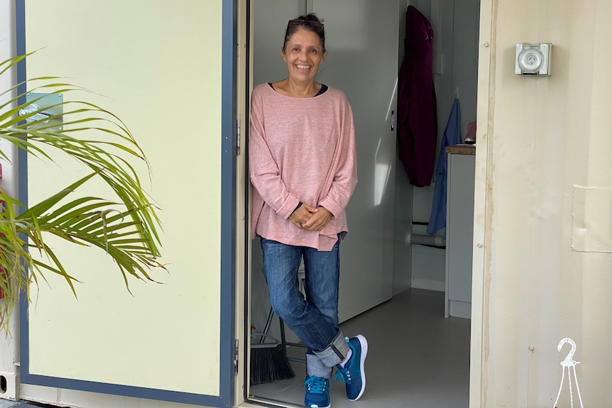 A woman stands in a doorway of a cream-coloured structure, grinning at the camera. A fern is off to the side.