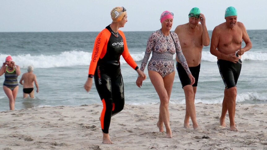Two mature aged women and two mature aged men walk on the beach out of the surf on a cold-looking day.