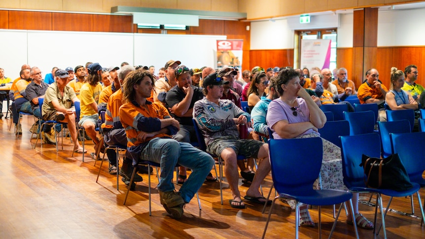 A group of people, mostly men, many in high-vis, listening to a speech in a community hall.