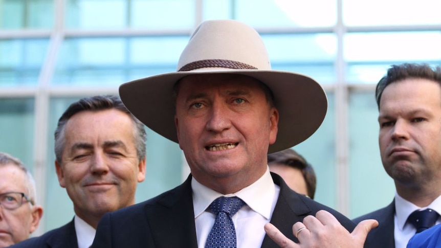 Barnaby Joyce wears an Akubra hat and does the hang loose hand gesture with his right hand while speaking to media.