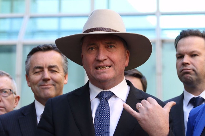 Barnaby Joyce wears an Akubra hat and does the hang loose hand gesture with his right hand while speaking to media.