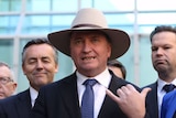 Barnaby Joyce wears an Akubra hat and does the hang loose hand gesture with his right hand while speaking to media.