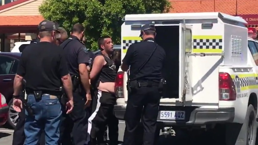A man in a singlet and black trackpants being pushed into a police vehicle