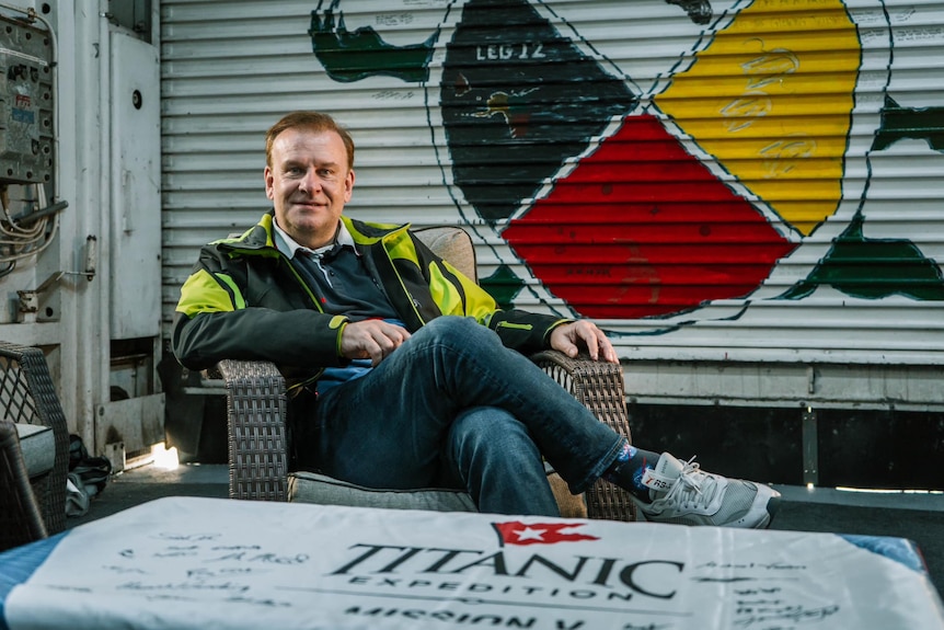 A middle-aged man in jeans and a jacket sits in a chair and smiles. In front is a table with Titanic Expedition written on it