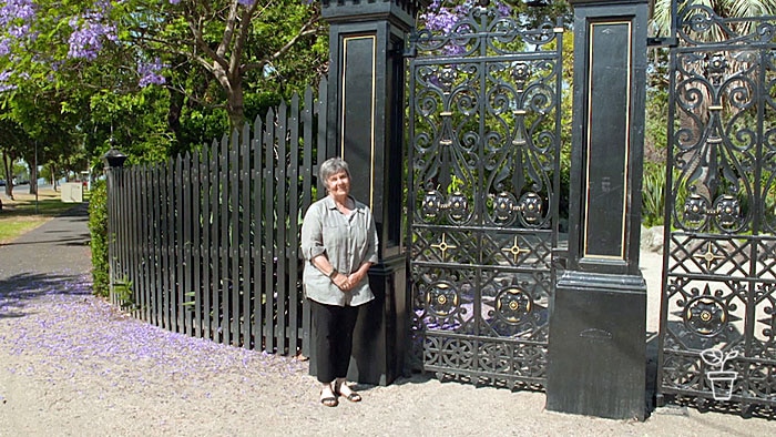 Woman standing outside large black metal gates and fence surrounding garden