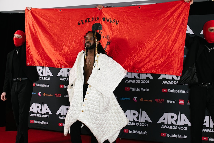 Genesis Owusu wearing a white gown, hair braided and standing on the red carpet with an eponymous sign behind him