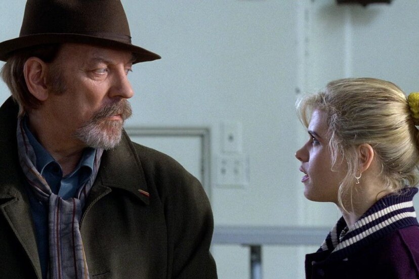 A man in a fedora and coat stands side on, making eye contact with a blonde teen girl in a varsity jacket.