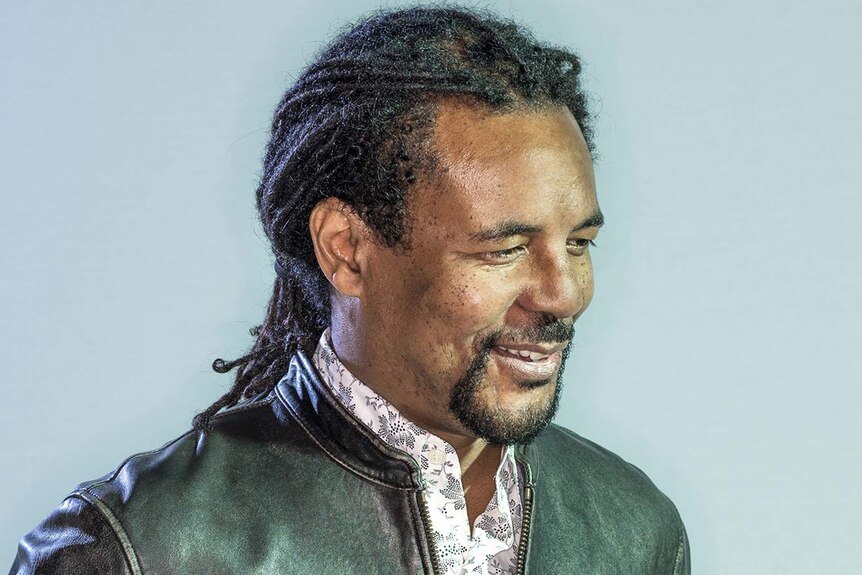 A man with long dreadlocks pulled into a pony tail and leather jacket  stands in front of a blue background and smiles.