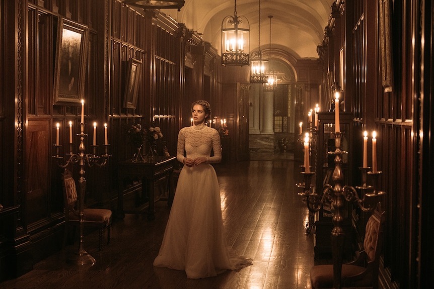 Samara Weaving looks at painting and walks down candlelit ornate Gothic style hallway in lace wedding dress and braided hair.