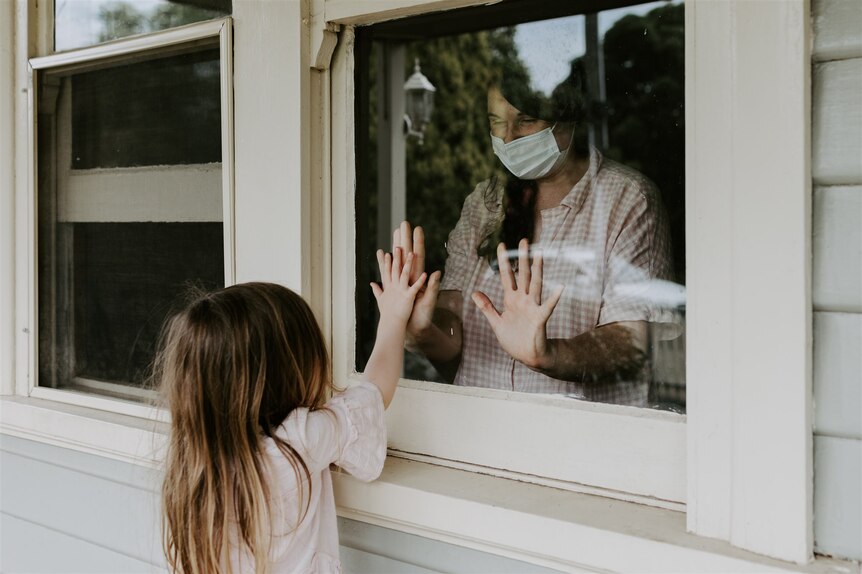 A small girl stands outside a window, holding her hand up to the glass. A masked woman inside does the same.