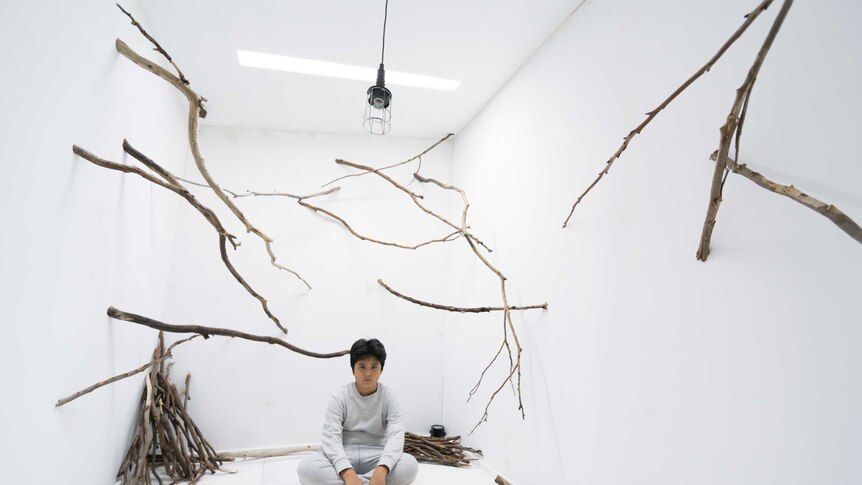 A young boy sits in a white room covered in sticks.