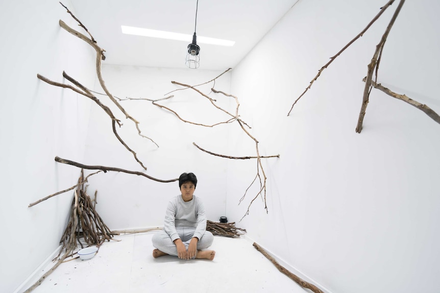 A young boy sits in a white room covered in sticks.