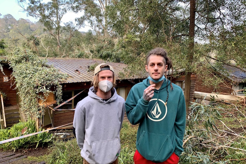 Two men stand in front of a damaged house marked with security tape and a fallen tree next to it.