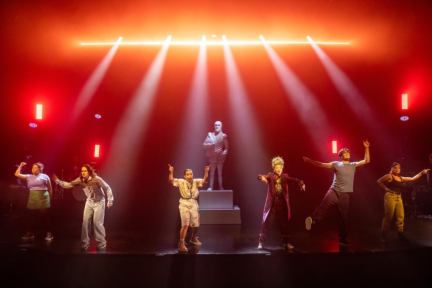 A cast of six performers sing out to the audience from a stage lit with red and orange backlights, with a statue in the centre.