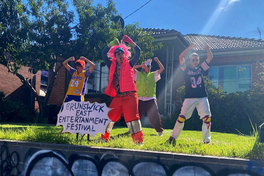 Four people dressed in flouro dance in front of a brick house in a suburban frontyard