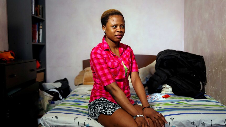 A young Nigerian woman with close cropped hair wearing a pink checked t-shirt sits on a bed with her hands on her knees.
