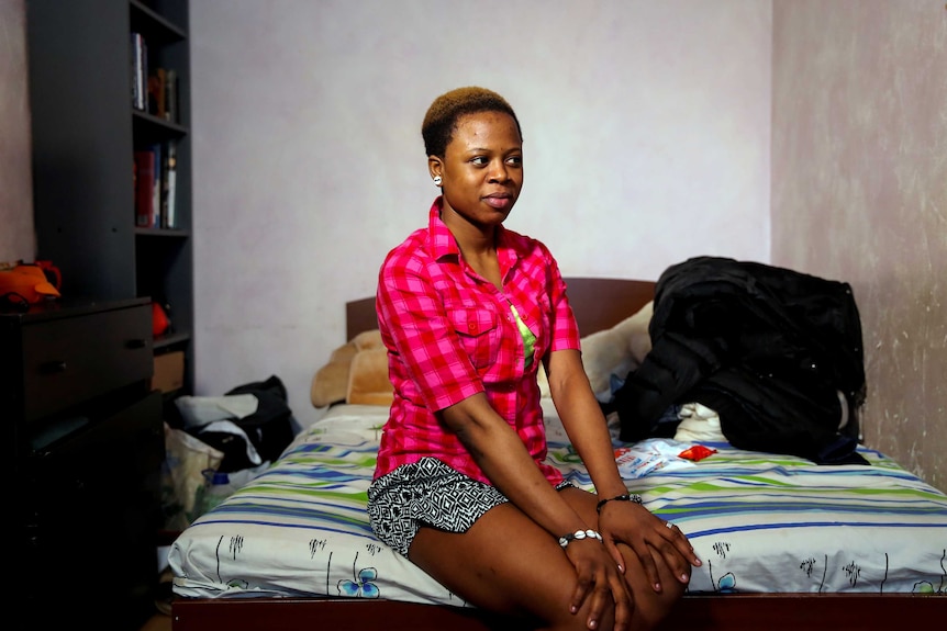 A young Nigerian woman with close cropped hair wearing a pink checked t-shirt sits on a bed with her hands on her knees.