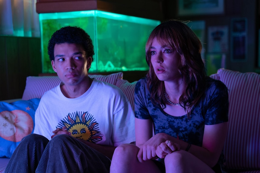 A film still of Justice Smith and Brigette Lundy-Paine, sitting together at night on a couch, their faces lit by a TV screen.