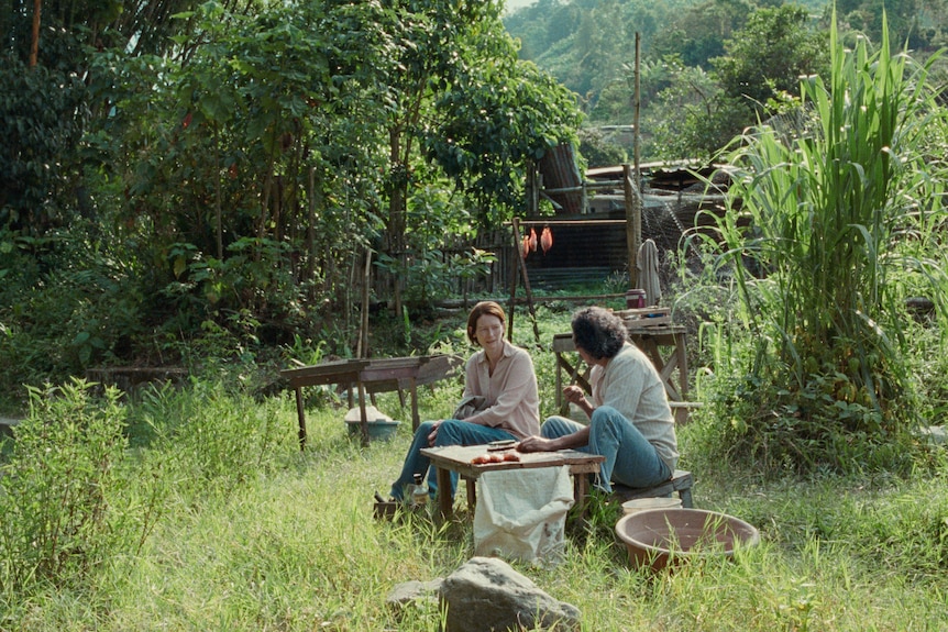 A middle-aged woman sits in an overgrown backyard in Colombia at a low table with a middle-aged man
