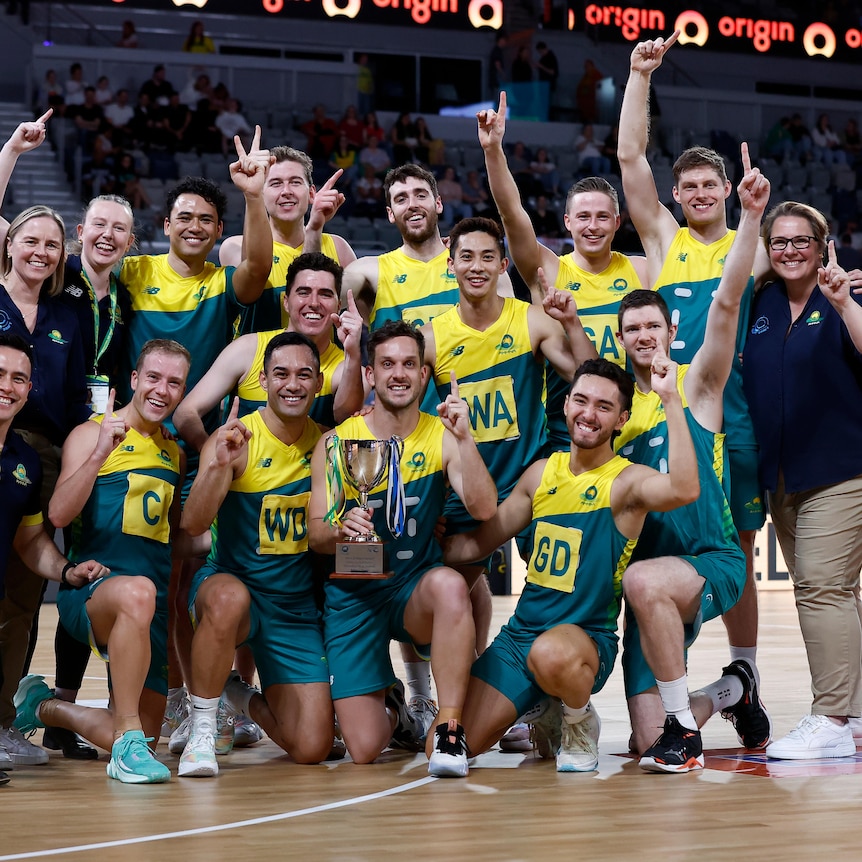 Men's netball players crowd together with coaching and support staff as they pose with the trophy